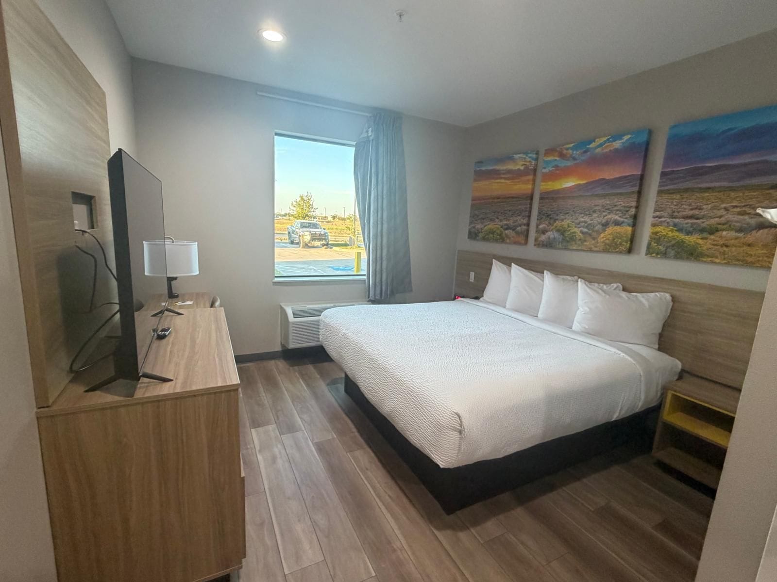 A hotel room with a large bed, a desk with a lamp, a flat-screen TV, and a window with a view of a parking lot.