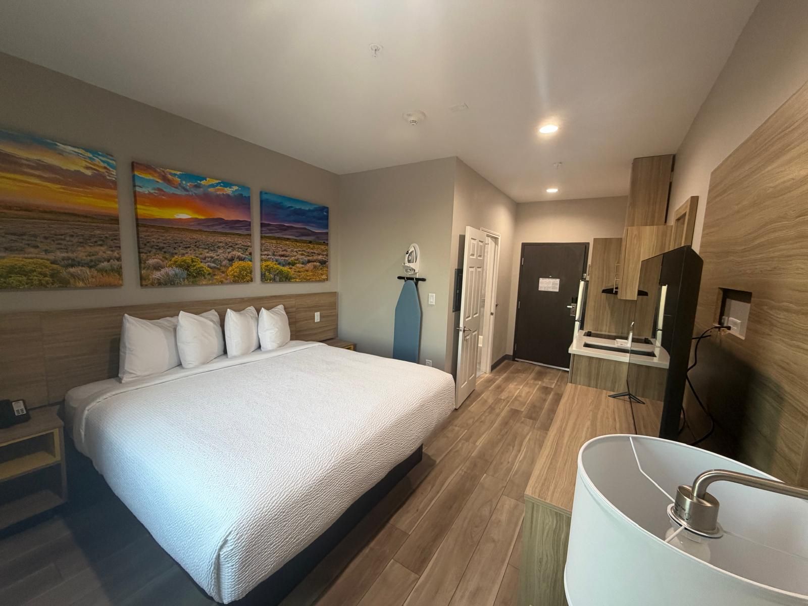 Habitación de hotel con una cama grande, un baño con lavamanos y espejo modernos, y tres cuadros de paisajes en la pared.