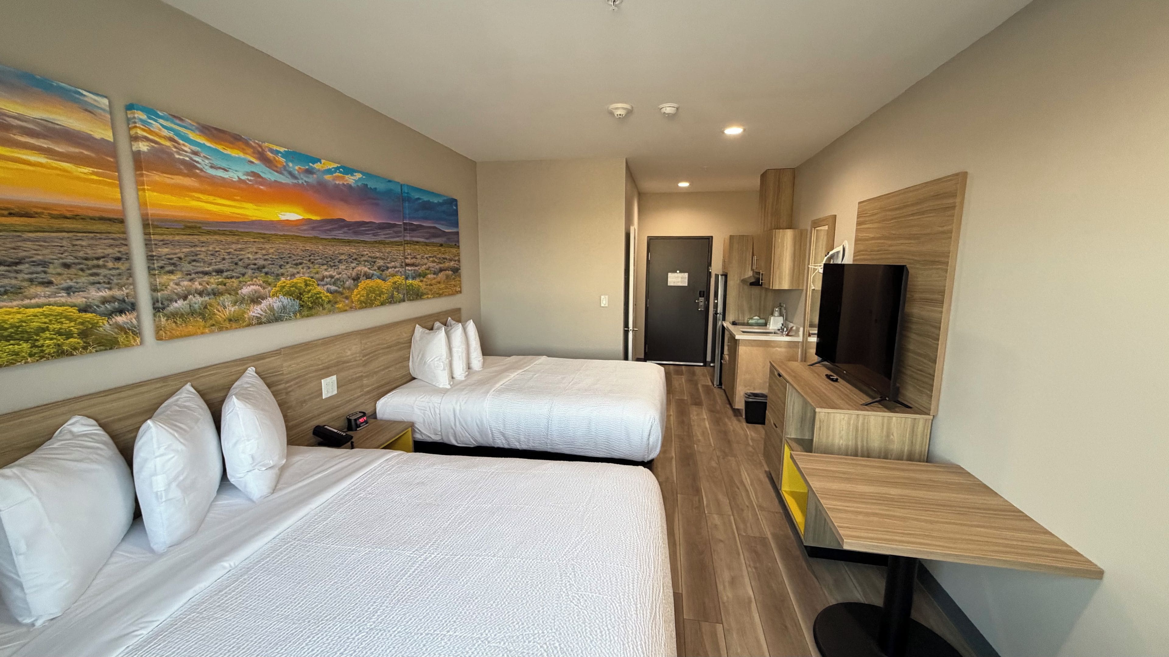 A hotel room with two beds, a TV, a desk, and a painting of a sunset.