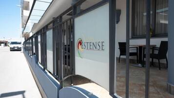 L’extérieur d’un hôtel avec une façade en verre, une enseigne indiquant « Hôtel Cestense » et des tables et chaises installées à l’extérieur.