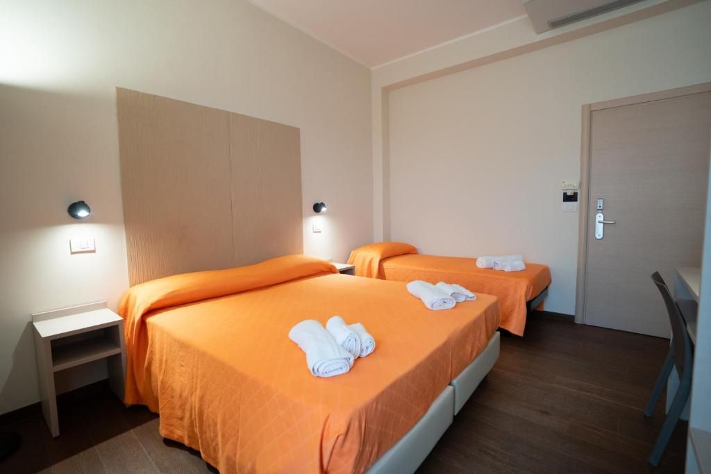 A hotel room with two beds, each with an orange bedspread and white towels. There is a desk with a chair, a door, and a light fixture on the wall.