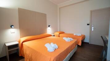 A hotel room with two beds, each with an orange bedspread and white towels. There is a desk with a chair, a door, and a light fixture on the wall.