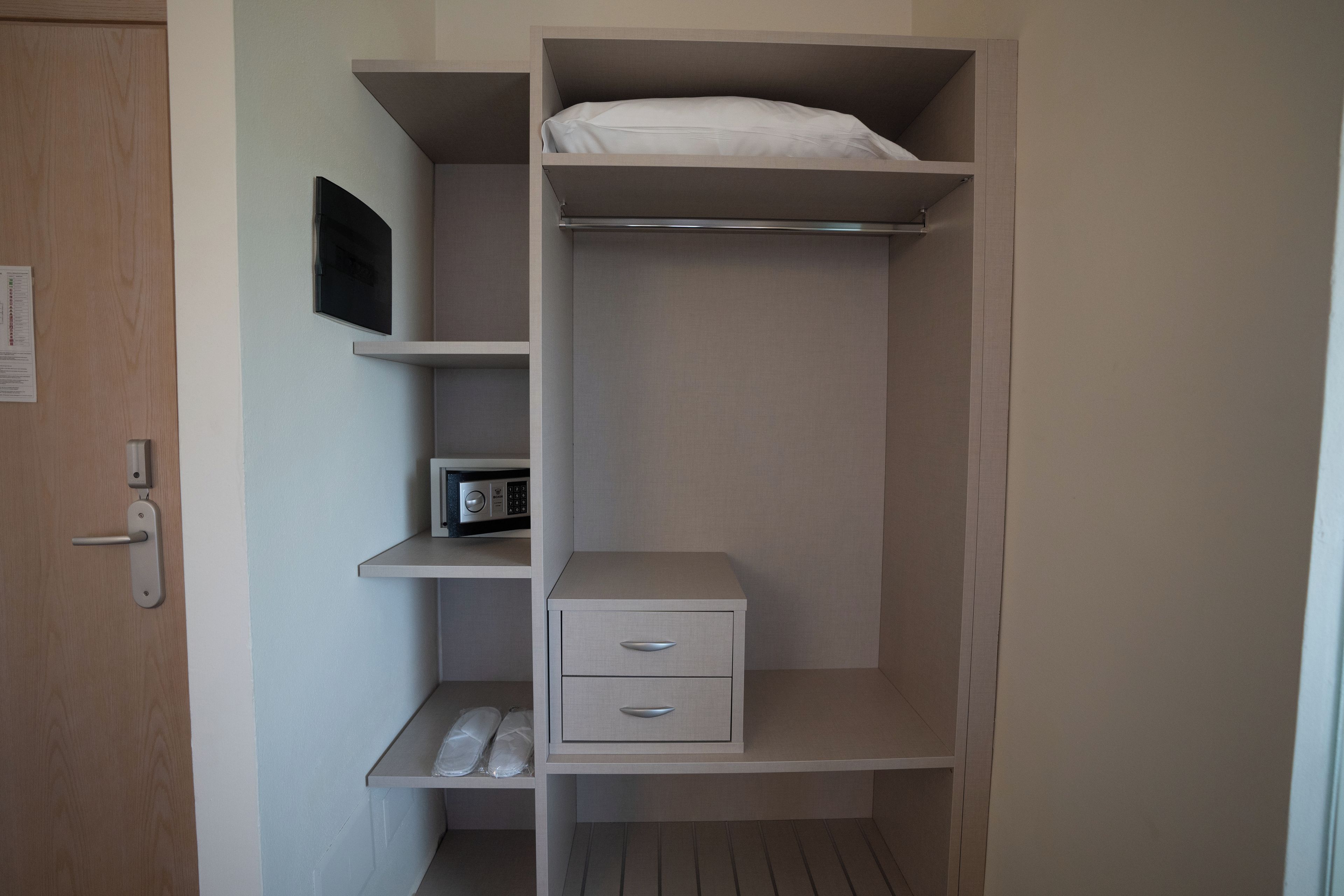 Ein kompakter, moderner Raum mit integriertem Kleiderschrank, der mehrere Regale und eine kleine Schublade bietet, sowie ein Bett mit weißer Matratze.