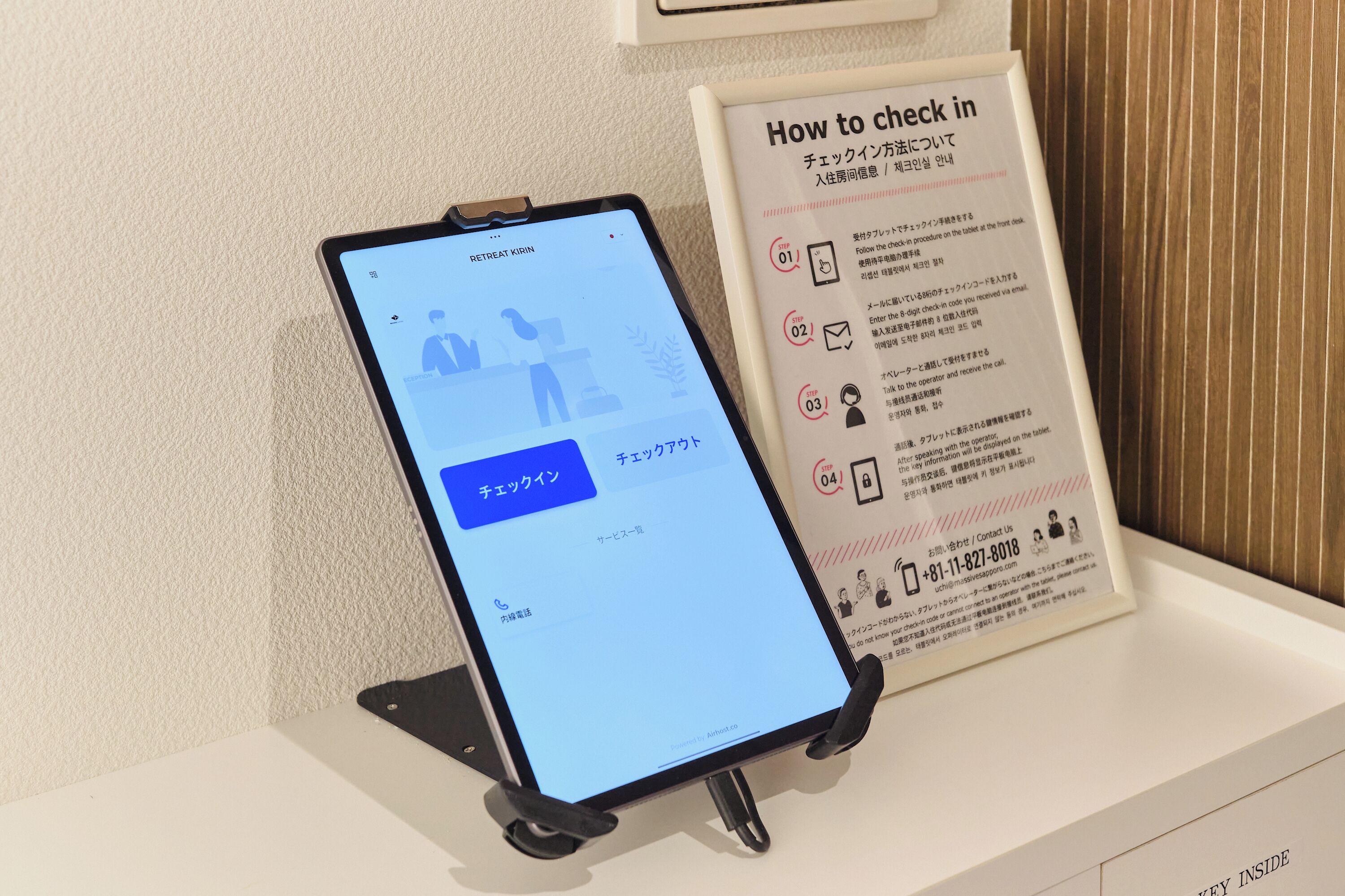 A tablet displaying a check-in process interface, a sign with check-in instructions, and a contact number.