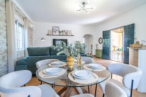 Ein modernes Wohnzimmer mit einem Esstisch aus Glas, gedeckt für vier Personen, umgeben von hellgrauen Stühlen. Ein grünes Sofa steht an der Wand, und darüber ist ein Flachbildfernseher angebracht.