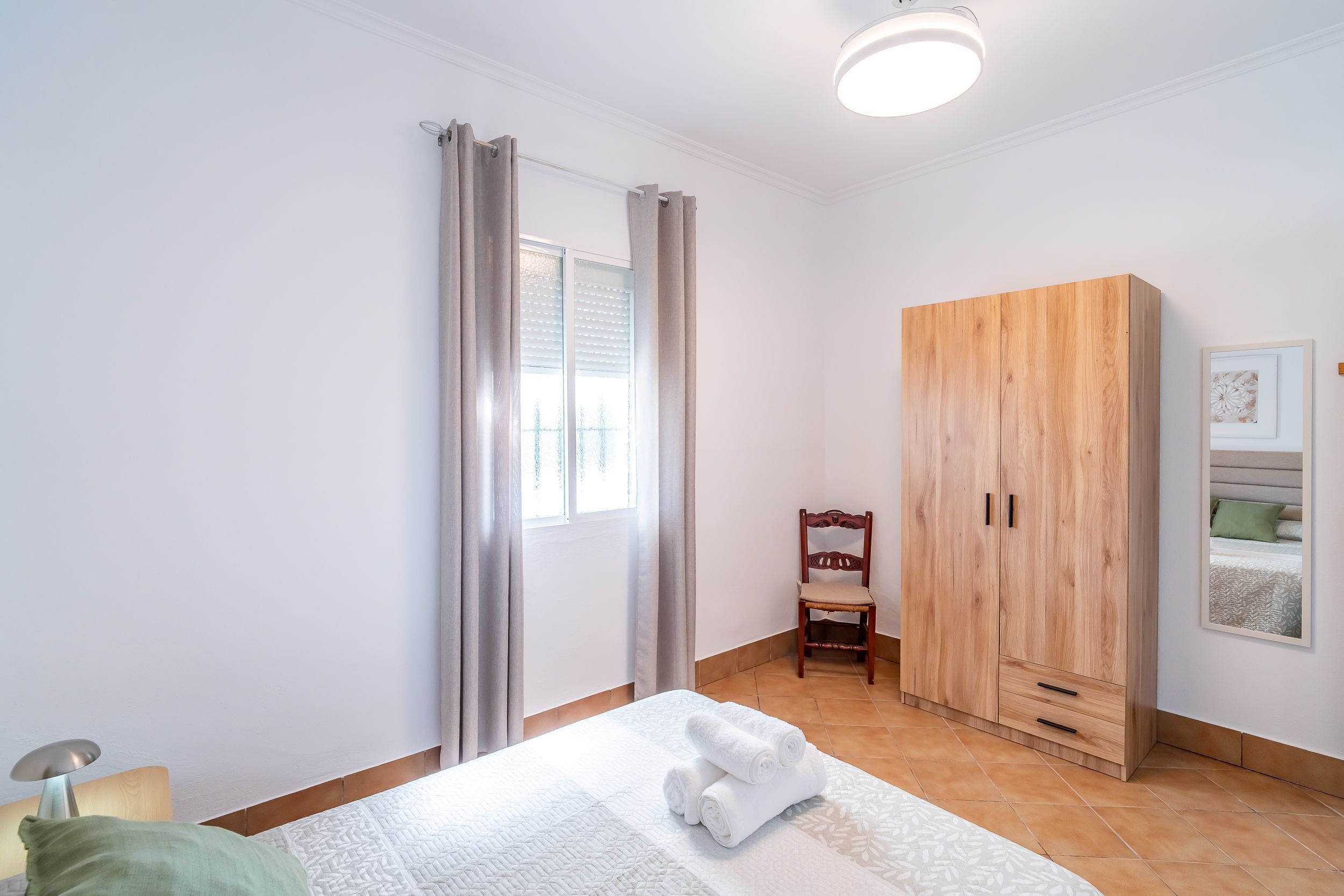 A bedroom with a bed, a wooden wardrobe, a chair, a window with curtains, and a wall-mounted picture.