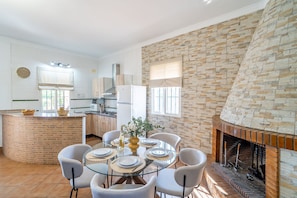 A modern kitchen with a brick island, a dining area with a glass table, and a stone fireplace. - Villa Manantial Holiday Home (La Puebla de Cazalla)