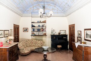 A room with a curved sofa, a round table, a bookshelf, a piano, and framed pictures on the walls.
