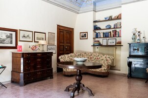 A room with a wooden dresser, a round table with a bowl, a floral sofa, a bookshelf, and a radiator.