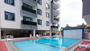 A swimming pool with lounge chairs, a covered car parking area, and a multi-story building with balconies. - Radium Residences View Palace Apartment (Mombasa)
