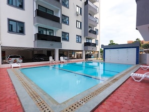 Una piscina con sillas de descanso, un estacionamiento techado para automóviles y un edificio de varios pisos con balcones.