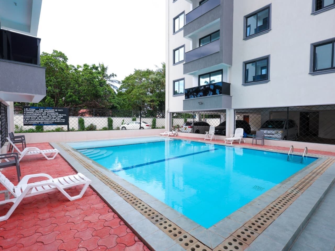 Une piscine avec des chaises longues, un espace délimité par une clôture et un immeuble à plusieurs étages doté de balcons.