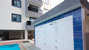 A swimming pool with a tiled deck, a covered area with multiple showerheads, and a building with balconies. - Radium Residences View Palace Apartment (Mombasa)