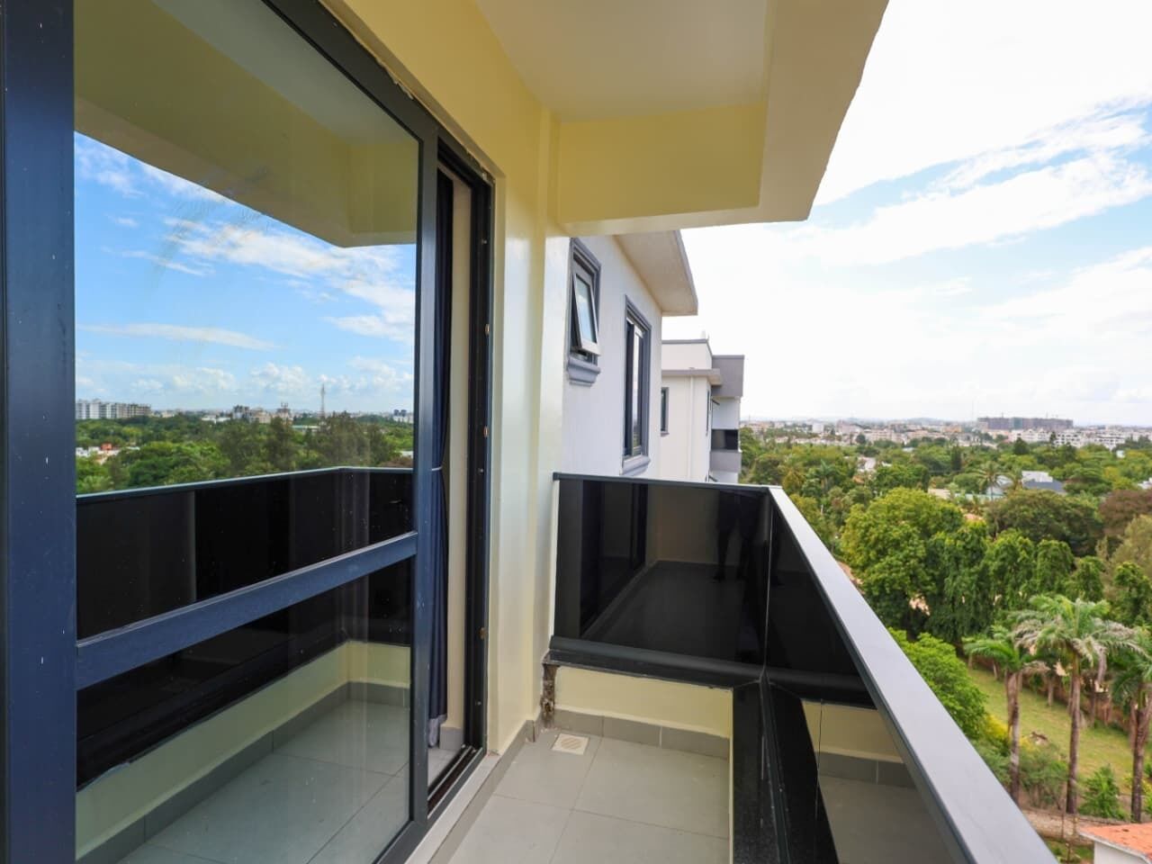 Een balkon met een glazen deur, een zwarte balustrade en uitzicht op een stadsbeeld en groen.