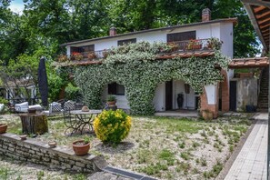 Ein Haus mit weißer Fassade, einem Balkon mit roten Ziegeln und einem Garten mit Blumentöpfen und einem gelben Busch.
