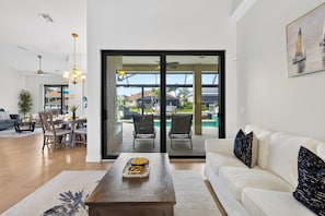 Ein Wohnzimmer mit einem weißen Sofa, einem Couchtisch mit einer Serviertafel voller Snacks, einem Essbereich mit Stühlen und einem Tisch und einer großen Schiebeglaswand, die zu einem Poolbereich führt.