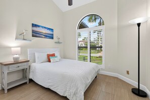Ein Schlafzimmer mit Bett, Nachttisch, Lampe, Kunstwerk, Fenster mit Jalousien und Blick ins Grüne.