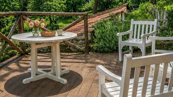 A wooden deck with a round table and two white chairs, surrounded by lush greenery and a clear blue sky.