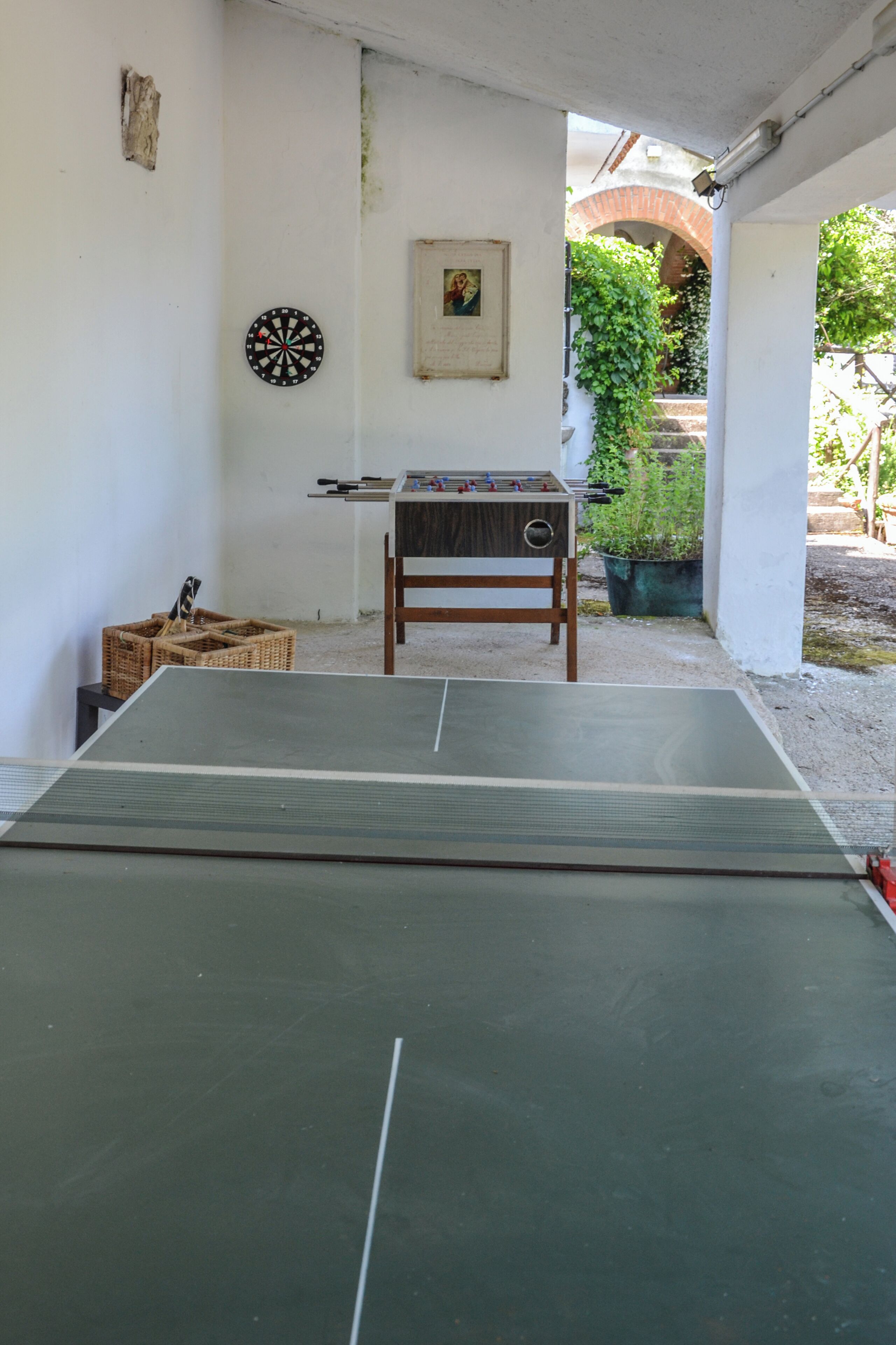 A ping pong table with a dartboard and foosball table in a room with white walls and a brick archway.