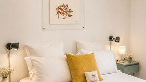 A neatly made bed with white and mustard pillows, a framed artwork above, and a bedside table with a lamp and a small plant. - Il Casale dei Castagni (Caprarola)