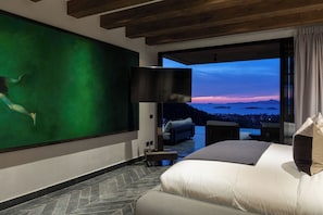 Una habitación de hotel moderna con una cama grande, un televisor de pantalla plana, un sofá y vistas a las montañas al atardecer.