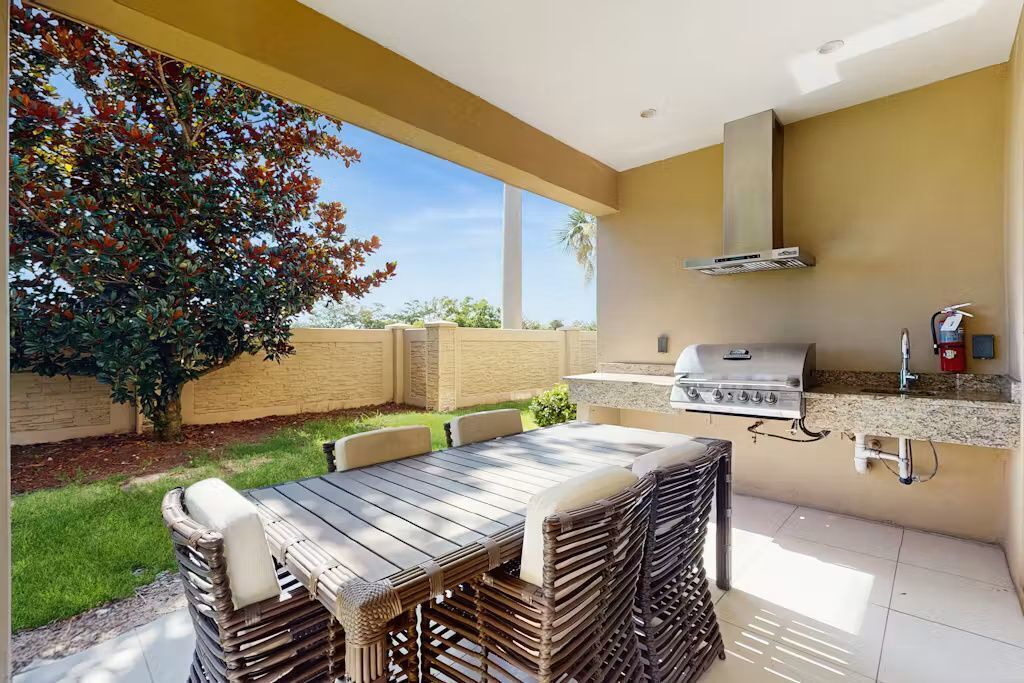 Una cocina exterior con una parrilla, una mesa de comedor con sillas de mimbre y vistas a un árbol y césped.