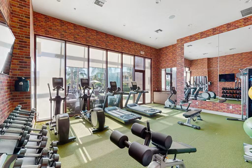 Un gimnasio con equipo de ejercicios, ventanas amplias y una pared de ladrillo.