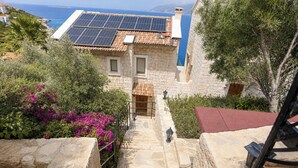 Edificio de piedra con una escalera que lleva a una puerta de madera, rodeado de vegetación y vista al mar.