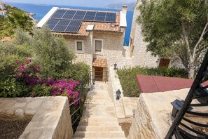 A stone building with a staircase leading to a wooden door, surrounded by greenery and a view of the sea. - Villa Ayışığı (Kaş)