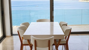Un comedor con mesa y sillas, ventanas amplias con vistas al mar y colinas.