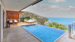 Una piscina infinita con vistas al mar y una terraza de madera con sillones reclinables.