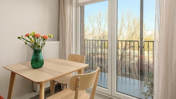 Ein Essbereich mit Tisch und Stühlen, einer Vase mit Blumen und einem großen Fenster mit Blick auf Bäume und Gebäude.