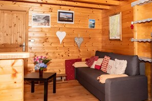 Interior de una cabaña de madera con un sofá, una mesa con un florero y cuadros en las paredes.