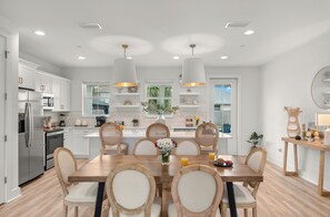 A modern kitchen with white cabinetry, a dining table with chairs, pendant lights, and a view of the outdoors. - Sleeps 26 • 2 Coastal Homes • 1 Block To Beach + Pool + Hot Tub | Twin Tides (Panama City Beach)