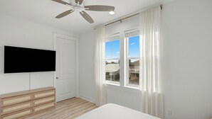 A bedroom with a bed, a ceiling fan, a TV mounted on the wall, and a window with curtains. - Sleeps 26 • 2 Coastal Homes • 1 Block To Beach + Pool + Hot Tub | Twin Tides (Panama City Beach)