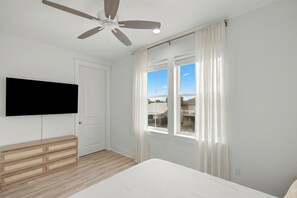 A bedroom with a bed, a ceiling fan, a TV mounted on the wall, and a window with curtains. - Sleeps 26 • 2 Coastal Homes • 1 Block To Beach + Pool + Hot Tub | Twin Tides (Panama City Beach)