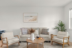 Ein Wohnzimmer mit einer Couch, zwei Sesseln, einem Couchtisch, einem gerahmten Bild, einer Vase, einem Weinglas und einer Obstplatte.