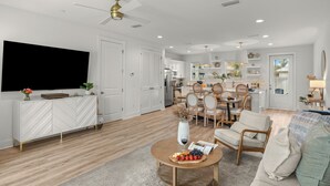 A modern living room with a flat-screen TV, a dining area with wooden chairs, and a kitchen in the background. - Sleeps 26 • 2 Coastal Homes • 1 Block To Beach + Pool + Hot Tub | Twin Tides (Panama City Beach)
