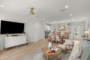 A modern living room with a flat-screen TV, a dining area with wooden chairs, and a kitchen in the background. - Sleeps 26 • 2 Coastal Homes • 1 Block To Beach + Pool + Hot Tub | Twin Tides (Panama City Beach)