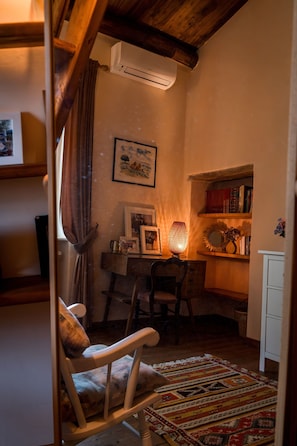 Ein gemütliches Zimmer mit Holzbalkendecke, einem Schreibtisch mit Lampe, einem Sessel, einem Bücherregal und einem gerahmten Bild an der Wand.
