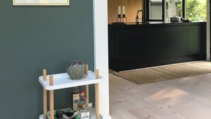 A room with a dark green wall, a framed artwork, a white cart with items, and a view of a kitchen area. - Architect-designed 80s house by Hornbæk (Hornbæk)
