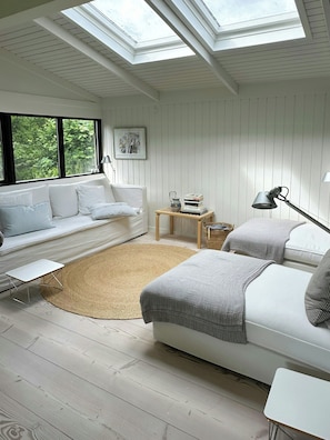 Ein modernes Schlafzimmer mit Dachfenster, weißen Wänden, einem weißen Sofa, einem runden Teppich und einem Nachttisch mit Büchern.