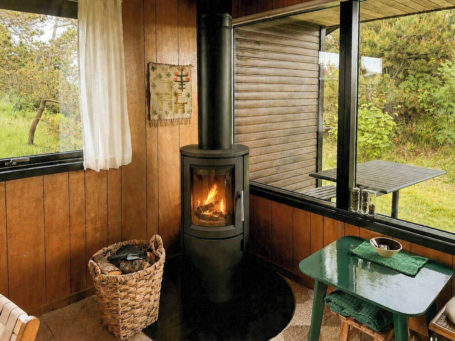 A cozy room with a wood-burning stove, a wicker basket, a green table, and a view of greenery outside.