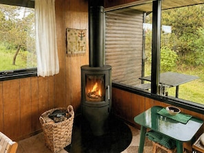 A cozy room with a wood-burning stove, a wicker basket, a green table, and a view of greenery outside. - Charming vintage house in North Sea nature (Ringkøbing)