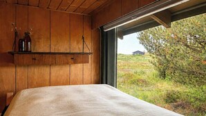 Ein Schlafzimmer mit Holzbalkendecke, einem Bett mit strukturierter Decke, einem Regal mit Gläsern und einem Fenster mit Blick auf eine grüne Landschaft.