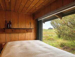 A bedroom with a wooden ceiling, a bed with a textured blanket, a shelf with jars, and a window with a view of a grassy landscape. - Charming vintage house in North Sea nature (Ringkøbing)