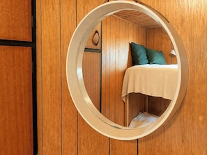 A circular mirror reflecting a bed with a green pillow and a white blanket, set against a wooden wall with a black trim. - Charming vintage house in North Sea nature (Ringkøbing)