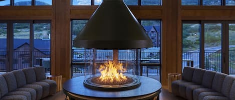 A modern interior with a central wood-burning stove, seating, and large windows offering views of a mountainous landscape.