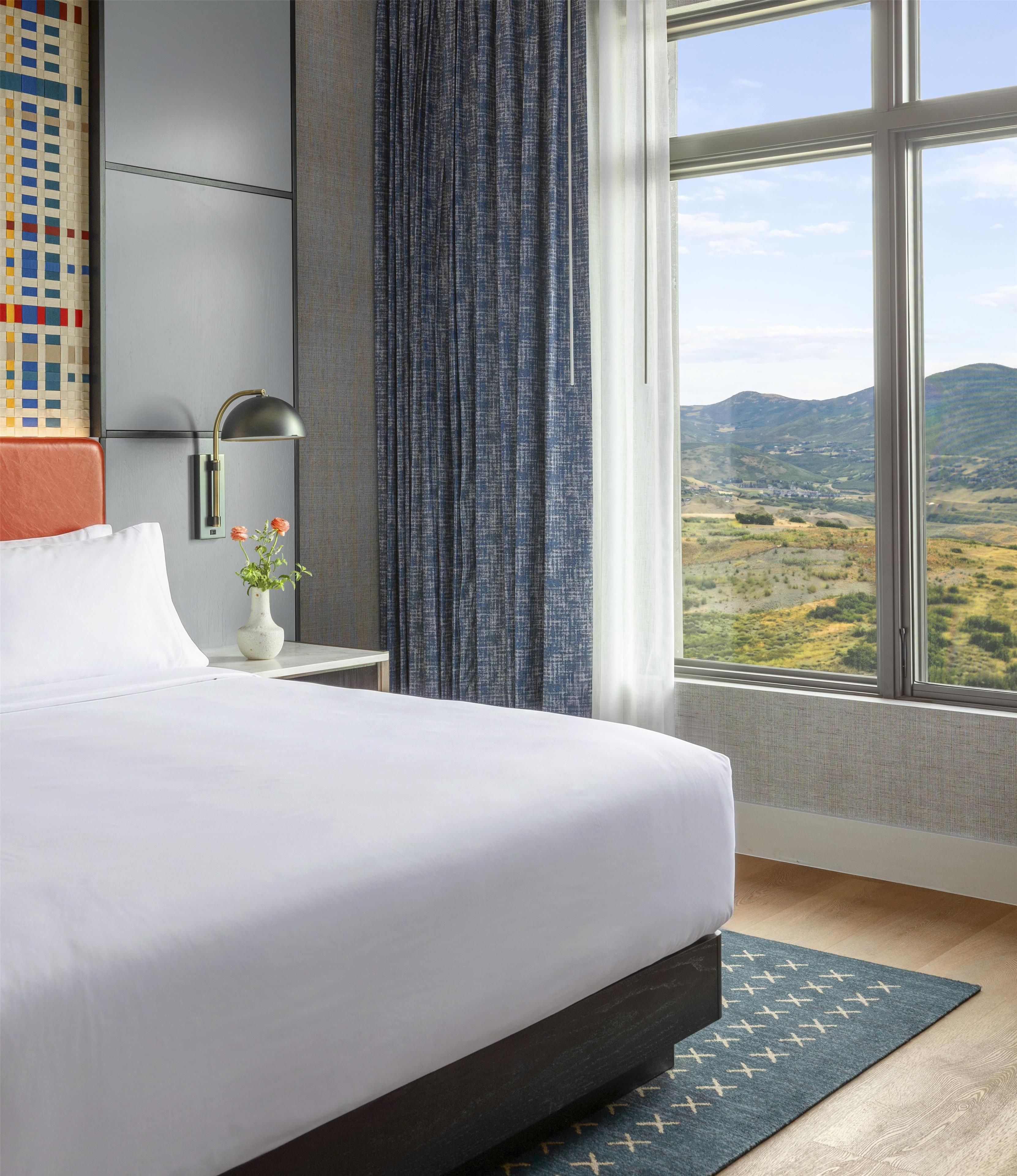 A hotel room with a large bed, a nightstand with a lamp, a vase with flowers, and a view of a landscape through a window.