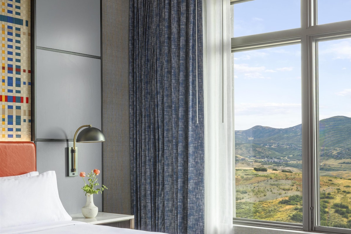 Una habitación de hotel con una cama grande, una mesita de noche con lámpara, un jarrón con flores y vistas al paisaje a través de una ventana.
